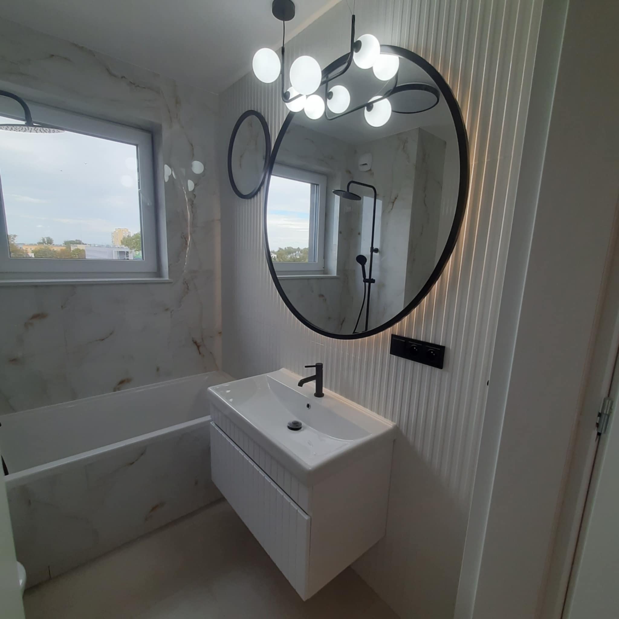 Nowoczesna łazienka z owalnym lustrem z podświetleniem LED, białą umywalką z szafką i wanną. Marmurowe płytki i dekoracyjne panele ścienne.