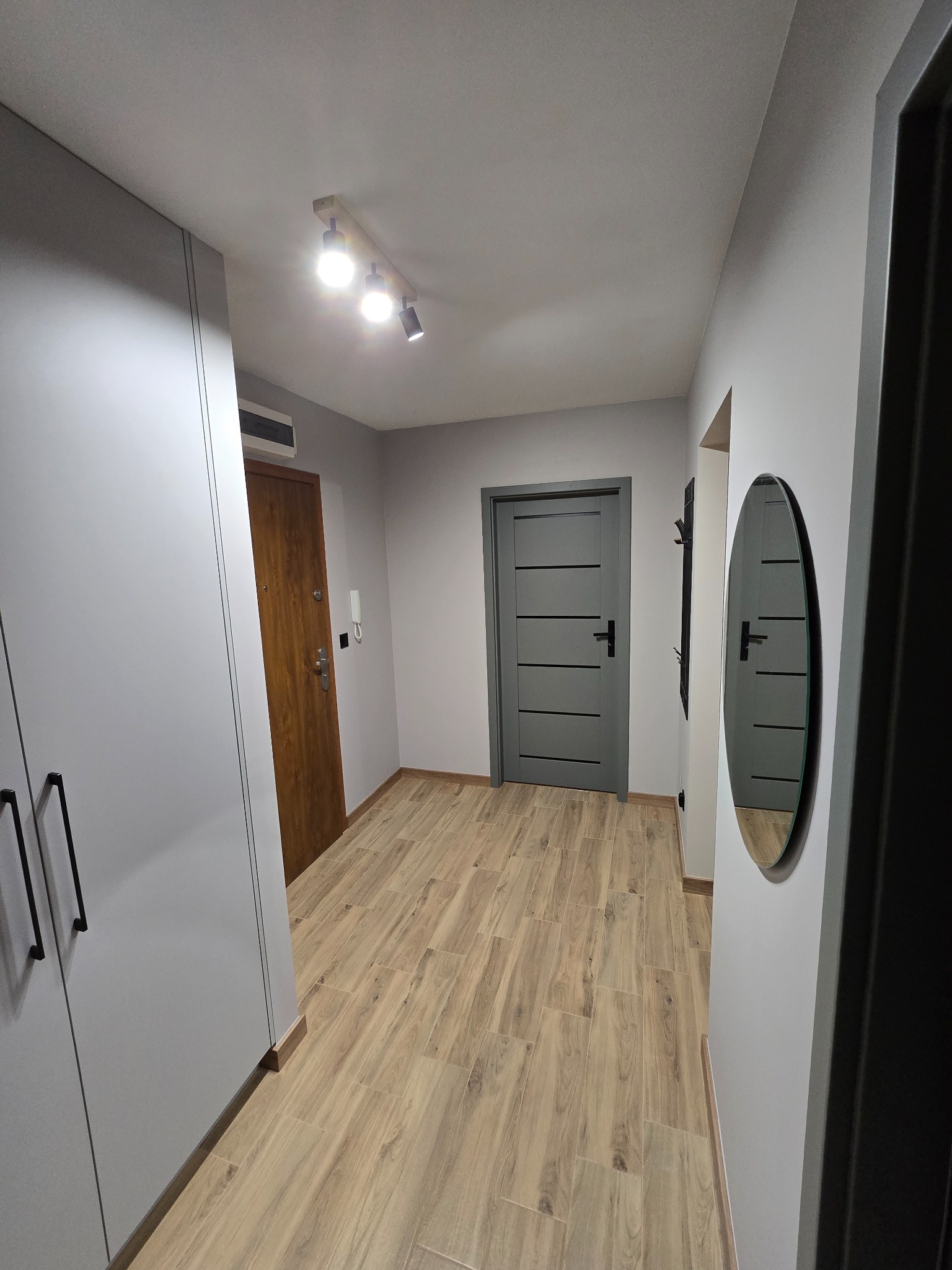 Nowoczesny przedpokój z drewnopodobną podłogą, szarą zabudową szafy i drzwiami w odcieniach szarości. Okrągłe lustro na ścianie. Oświetlenie sufitowe.