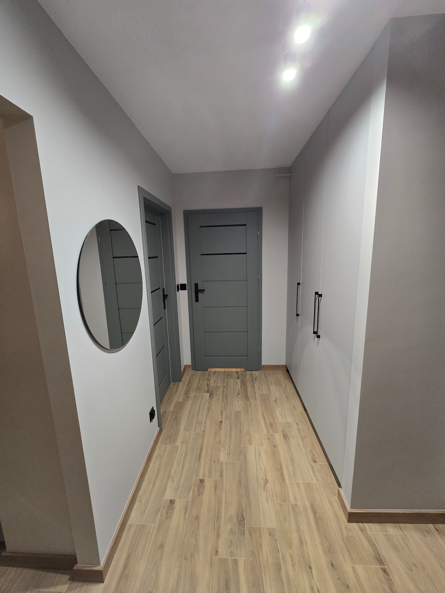 Nowoczesny przedpokój w szarościach: drewnopodobna podłoga, szare drzwi z poziomymi frezami, okrągłe lustro i szafa w zabudowie z czarnymi uchwytami.