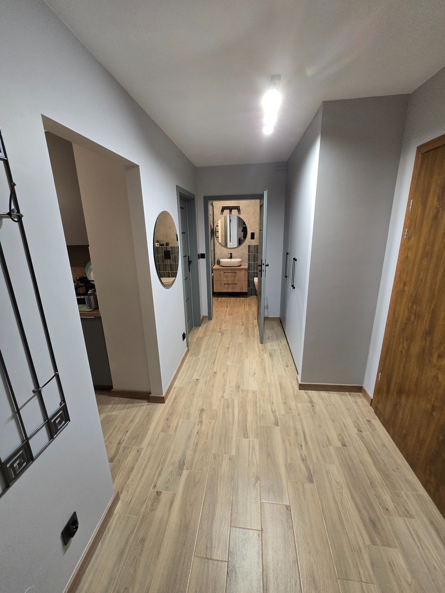 Nowoczesny korytarz z drewnopodobną podłogą, szarą kolorystyką ścian i widokiem na łazienkę z okrągłym lustrem. Minimalistyczny design, oświetlenie sufitowe.