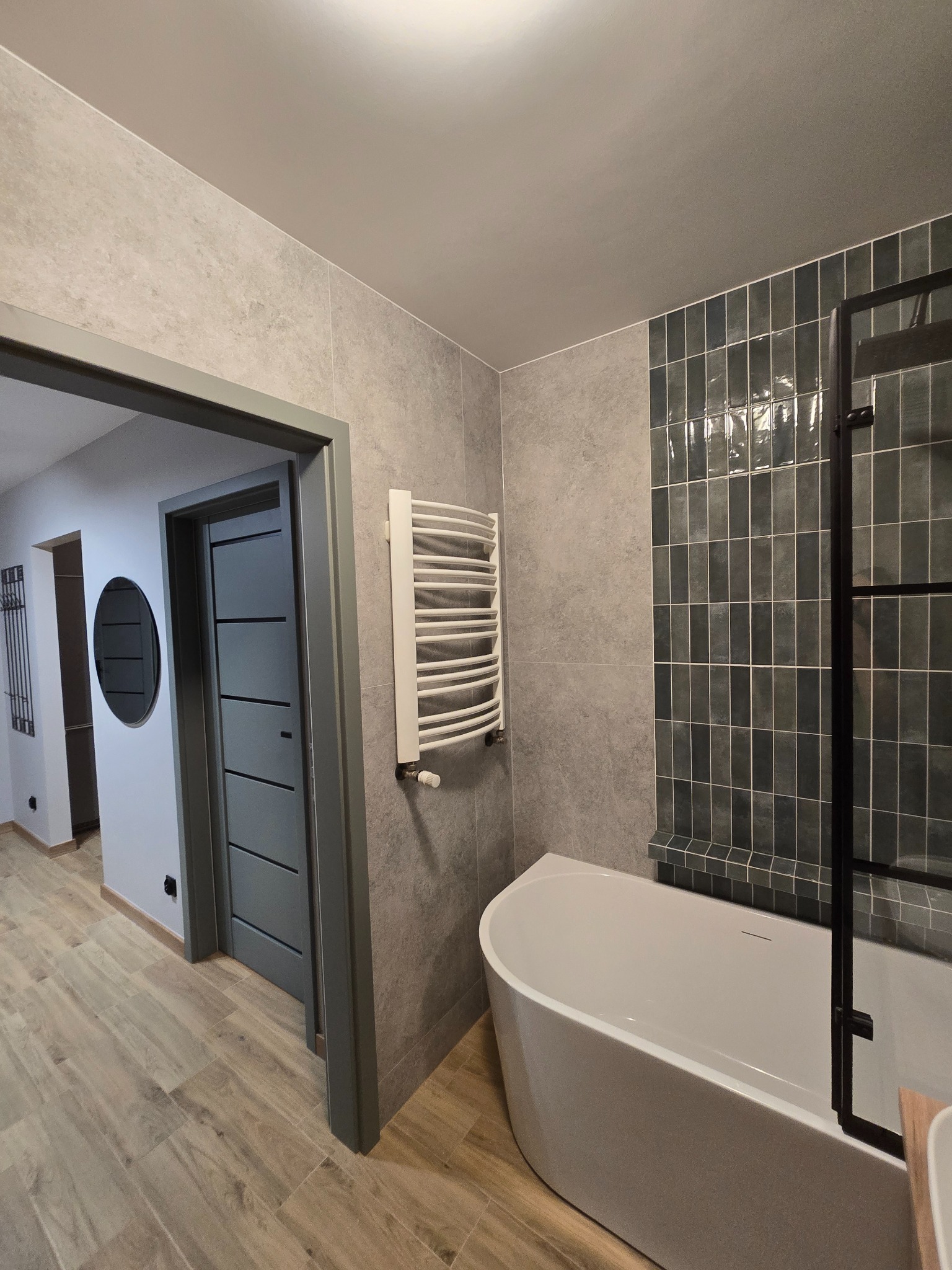 Nowoczesna łazienka po remoncie: szare płytki, wanna z parawanem, grzejnik drabinkowy i drzwi w odcieniach szarości. Widok z perspektywy wejścia.