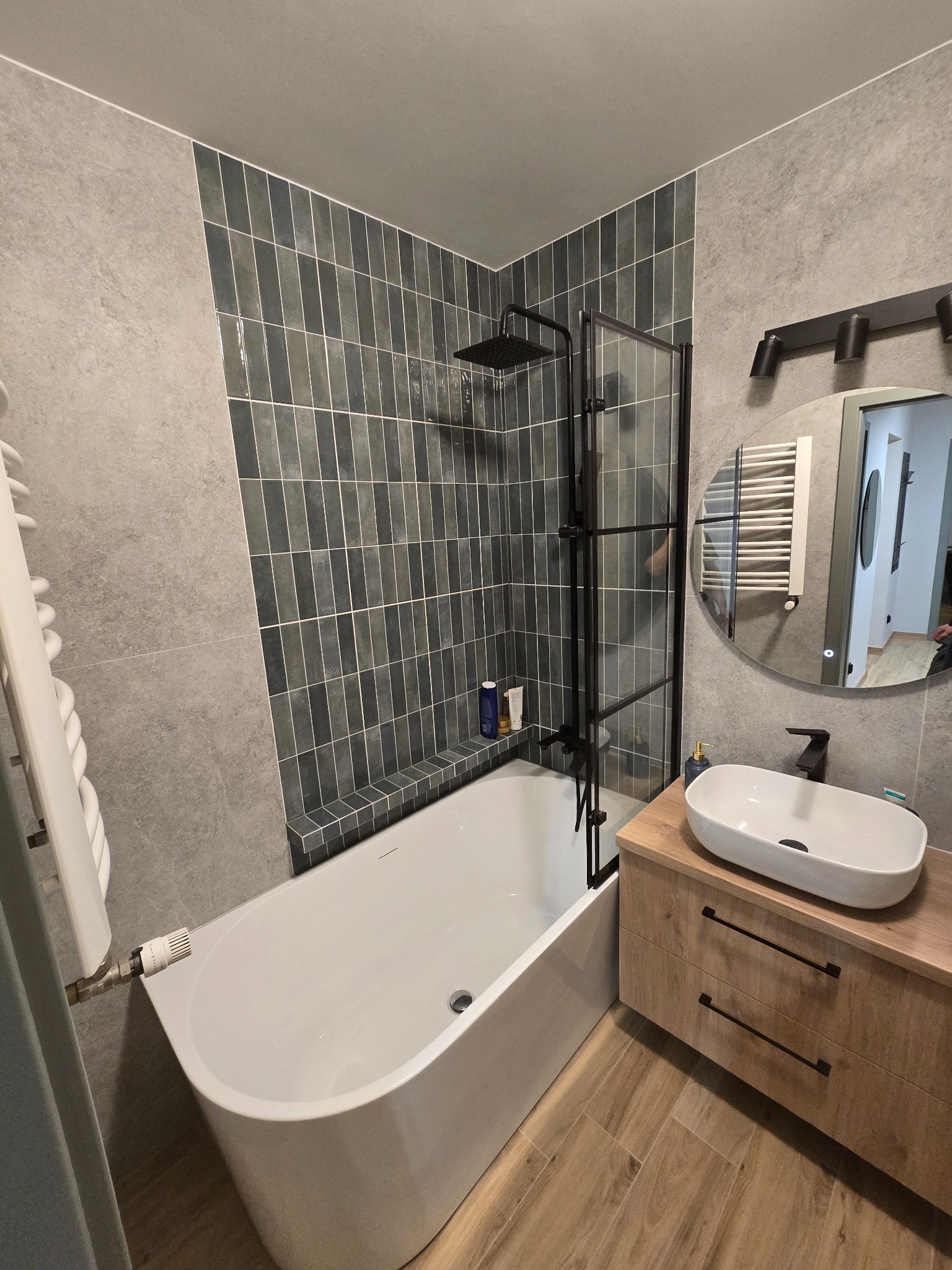 Nowoczesna łazienka z wanną, kabiną prysznicową typu walk-in z czarnymi ramami, umywalką nablatową na drewnianej szafce i szarymi płytkami na ścianach.