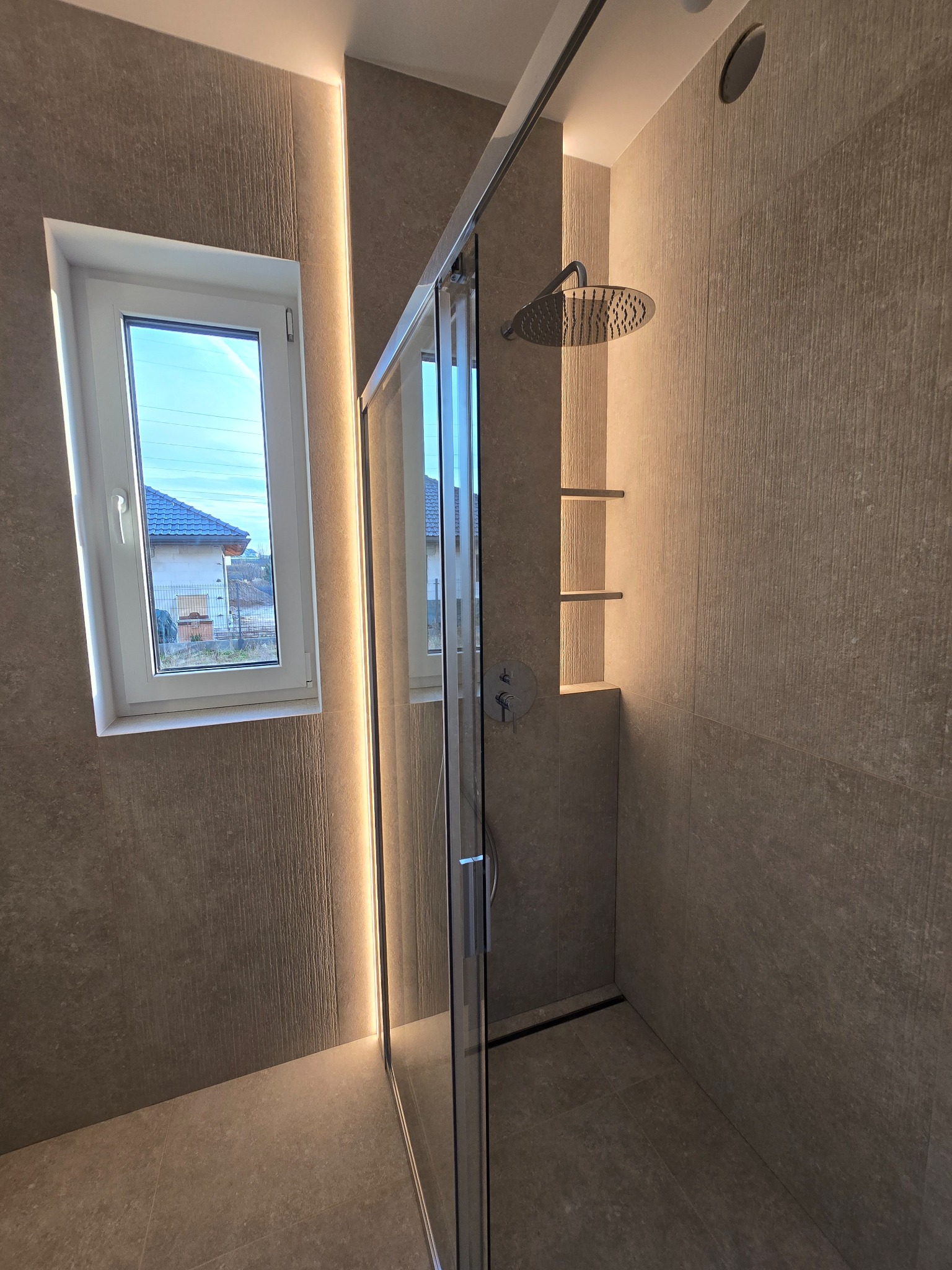 Nowoczesna kabina prysznicowa z deszczownicą i wnęką z półkami, podświetlona listwami LED. Widok na okno z widokiem na dom. Beżowe płytki na ścianach i podłodze.