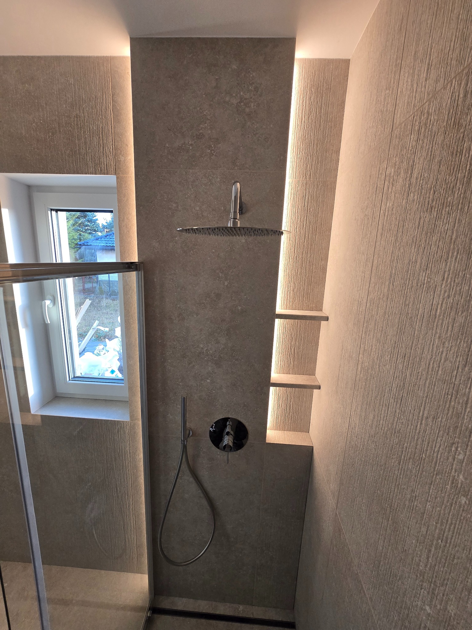 Nowoczesna kabina prysznicowa z deszczownicą i półkami wnękowymi, podświetlona listwami LED. Minimalistyczny design z szarymi płytkami o wyraźnej fakturze.