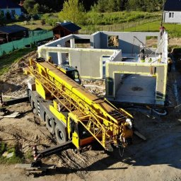 TECTA PROSTA SPÓŁKA AKCYJNA - Żółty dźwig budowlany w trakcie montażu ścian domu z prefabrykatów żelbetowych na fundamencie, widoczni pracownicy na budowie, otoczenie: teren budowy z domami w tle.