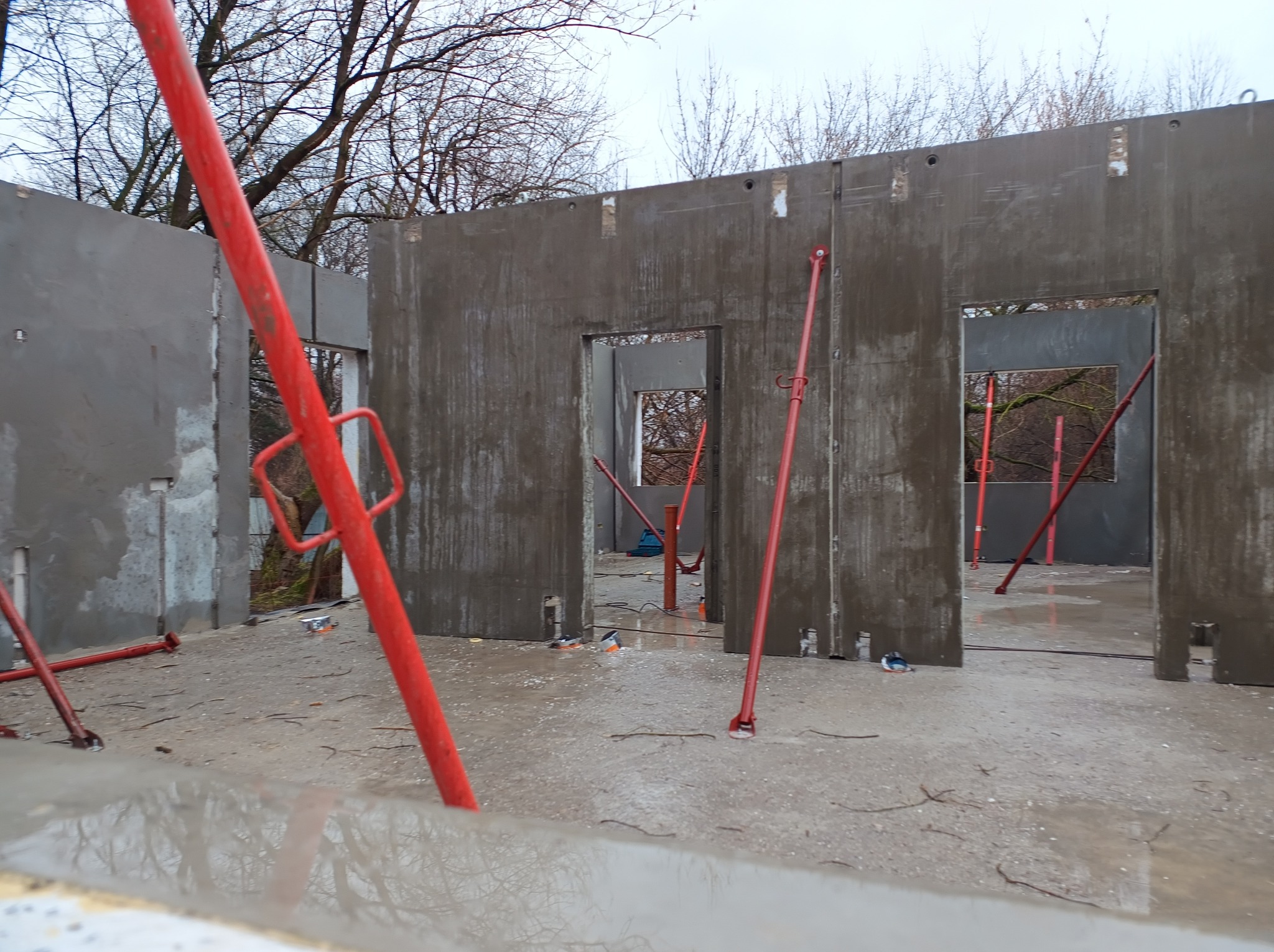 Wnętrze budynku w trakcie budowy z prefabrykatów betonowych, widoczne czerwone podpory, surowy beton i otwarte przestrzenie okienne.