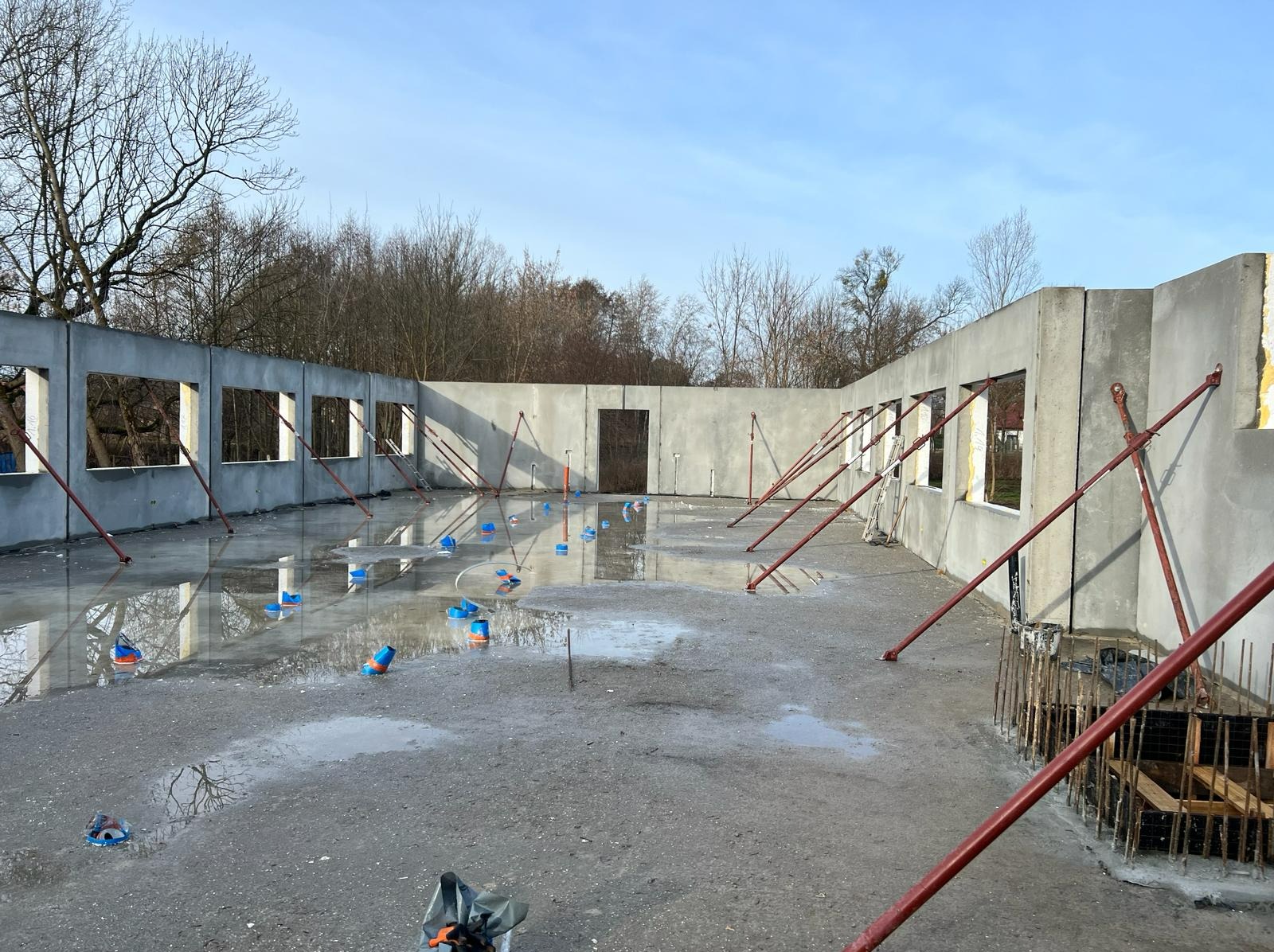 Wnętrze budynku w trakcie budowy z żelbetowymi ścianami, widoczne podpory i mokra posadzka, odbijająca niebo i drzewa za oknami.