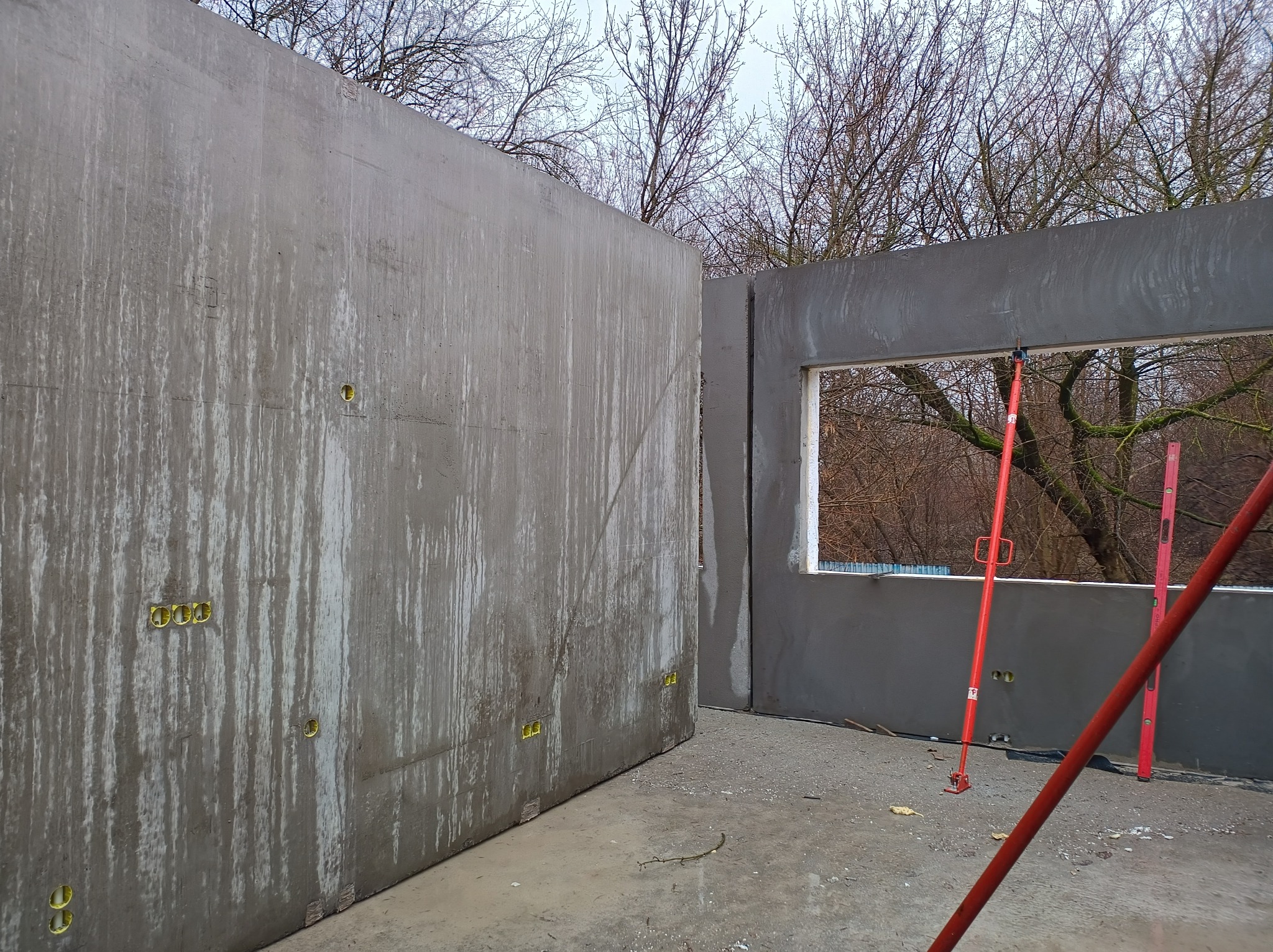 Surowe ściany z betonu z widocznymi puszkami elektrycznymi i podporami teleskopowymi, w tle drzewa bez liści.