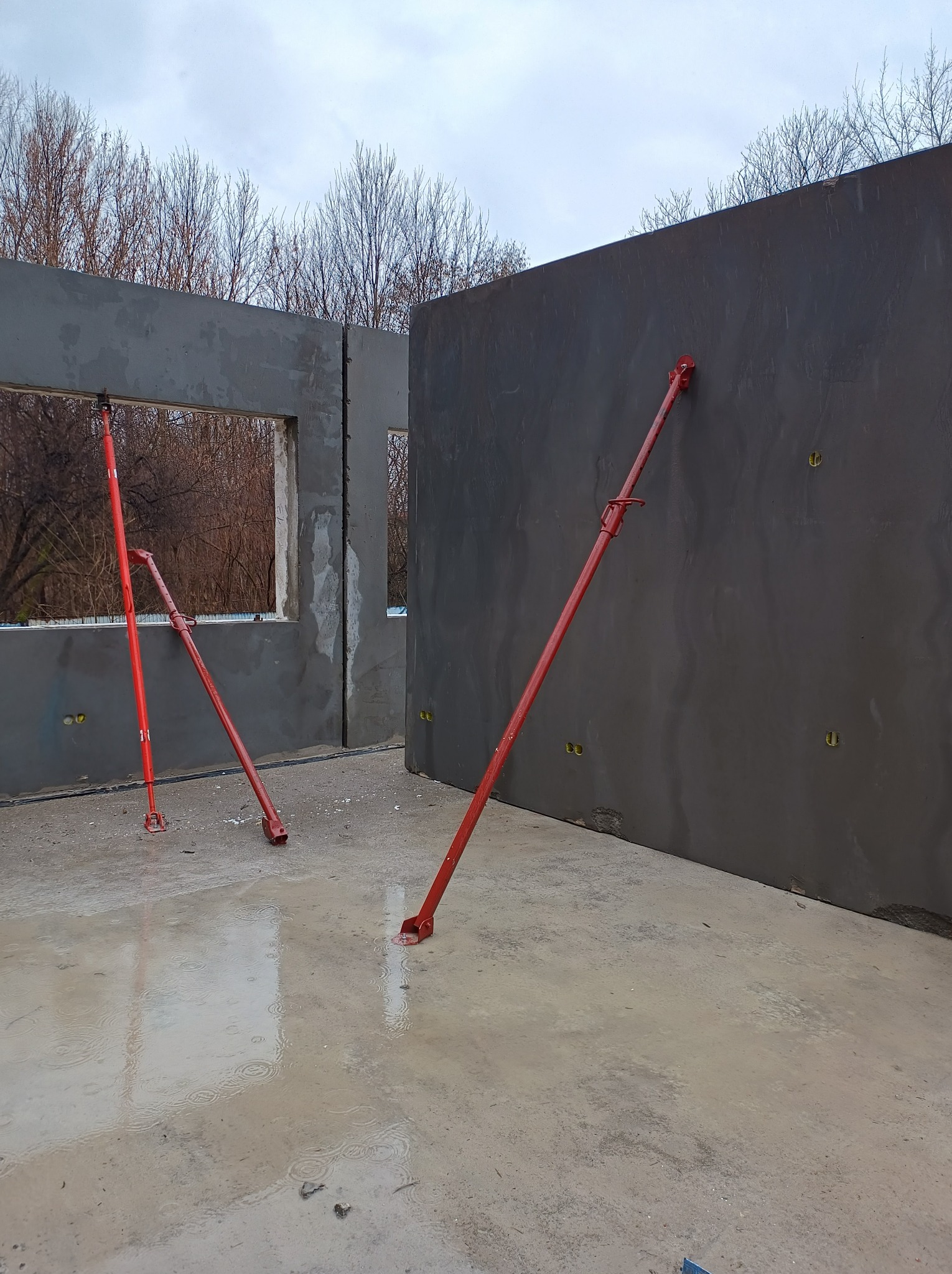 Wznoszenie ścian żelbetowych z otworami okiennymi, wspartych czerwonymi stemplami budowlanymi na mokrej, betonowej posadzce. W tle widoczne drzewa bez liści.