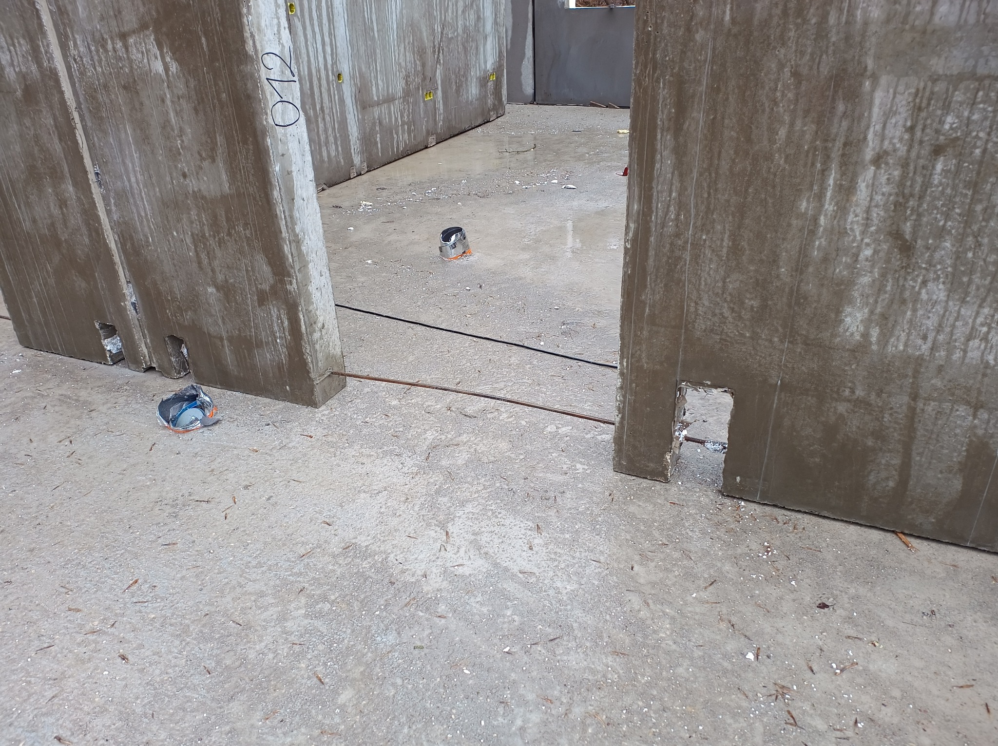 Surowy stan budowy: Wnętrze wznoszonego budynku z prefabrykatów betonowych, widoczne ściany z otworami montażowymi, betonowa posadzka z resztkami materiałów budowlanych i puszką po farbie.