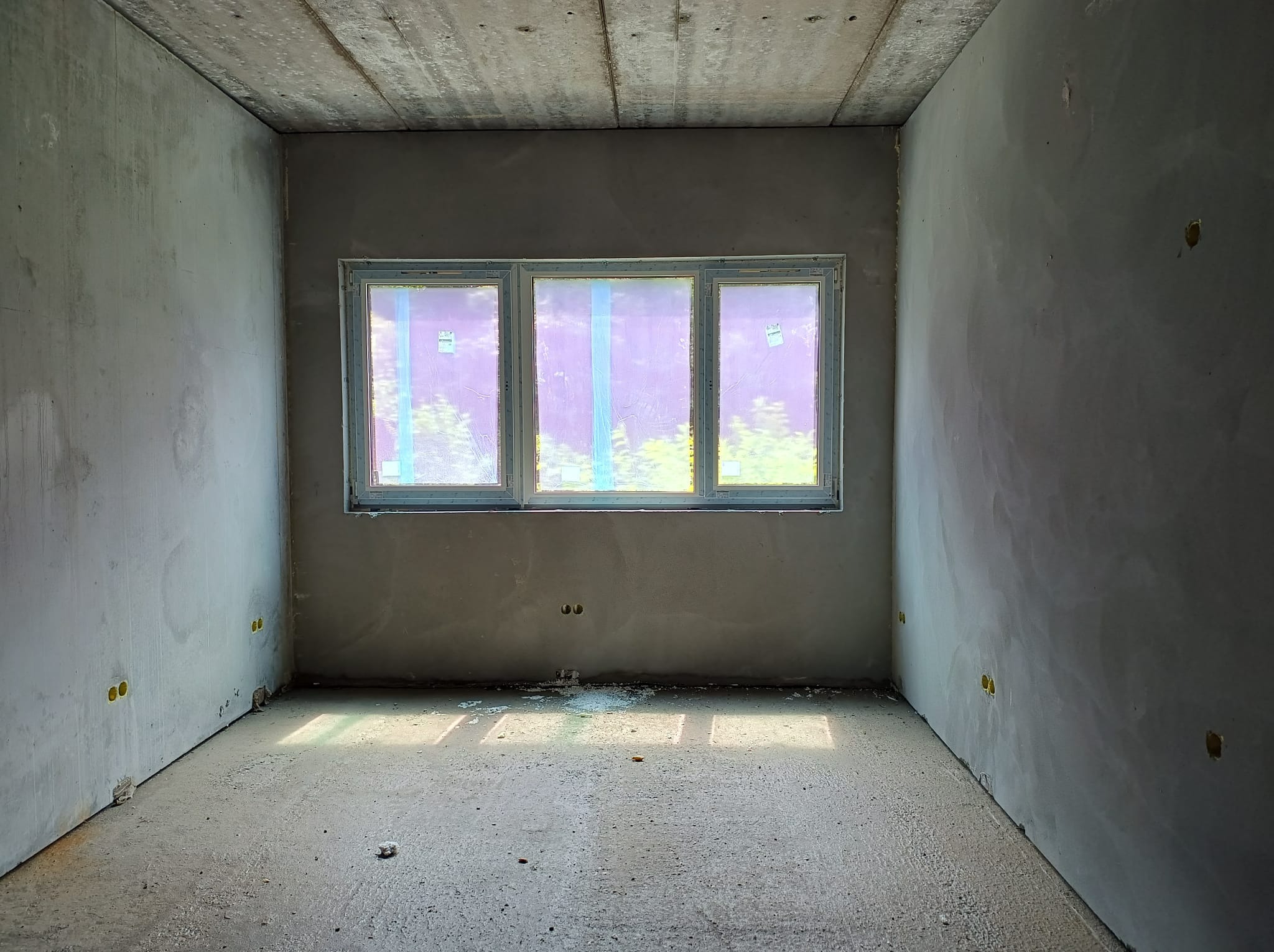 Surowe wnętrze budynku w stanie deweloperskim z trzema oknami zabezpieczonymi folią, widoczne puszki elektryczne w ścianach i betonowa podłoga.