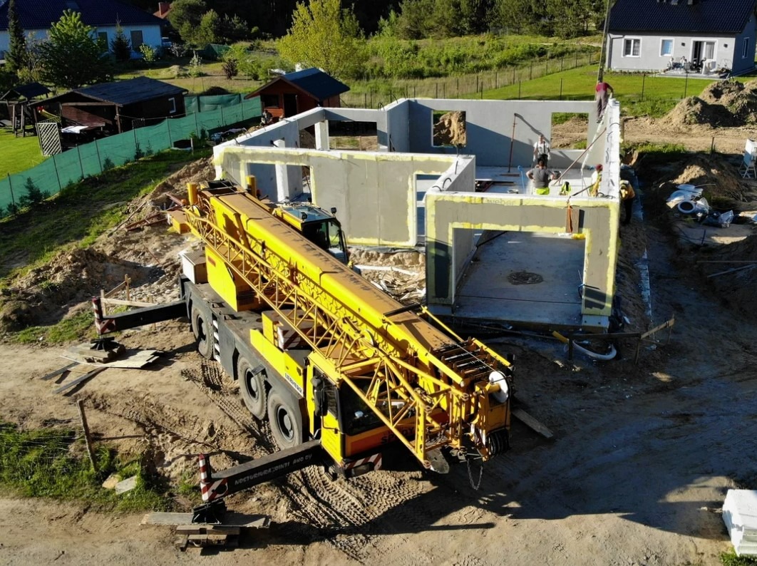 Żółty dźwig budowlany w trakcie montażu ścian domu z prefabrykatów żelbetowych na fundamencie, widoczni pracownicy na budowie, otoczenie: teren budowy z domami w tle.