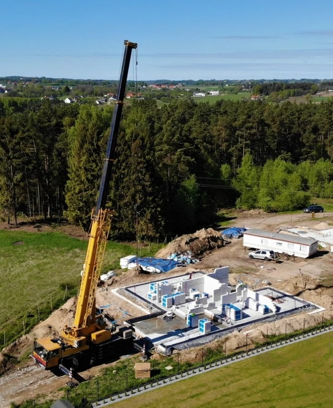 Widok z lotu ptaka na plac budowy z żółtym dźwigiem, wznoszone ściany z prefabrykatów na betonowej płycie fundamentowej, barakowóz i zalesione wzgórza w tle.