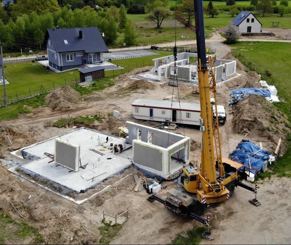 Montaż ścian domu modułowego z użyciem dźwigu na przygotowanym fundamencie, widoczny przyczepa mieszkalna i dom w tle.