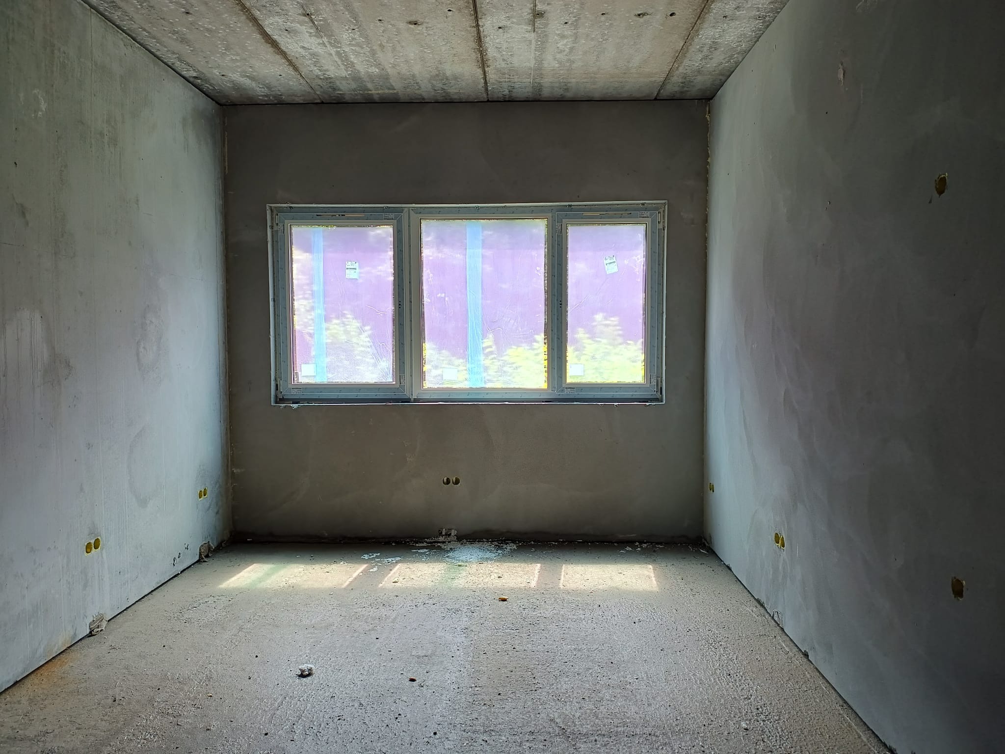 Surowe wnętrze budynku w stanie deweloperskim z trzema oknami zabezpieczonymi folią, widoczne ślady po instalacji elektrycznej w ścianach i surowy beton na podłodze i suficie.
