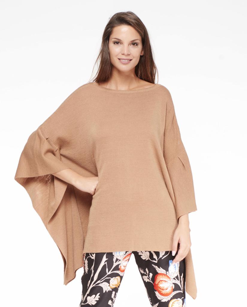 Kobieta prezentuje beżowy, dzianinowy ponczo-sweter z rękawami 3/4 i spodnie w kwiatowy wzór na białym tle.