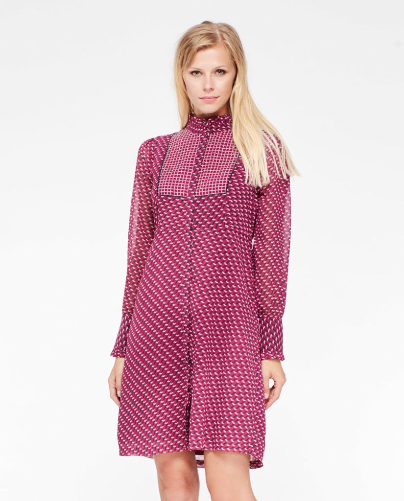 Modelka prezentuje zwiewną sukienkę z geometrycznym wzorem w odcieniach fuksji i bieli, z wysokim kołnierzykiem i bufkami przy mankietach, na jednolitym białym tle.