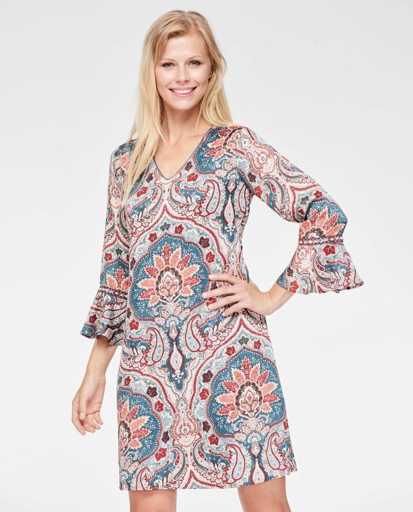 Uśmiechnięta blondynka prezentuje sukienkę w etniczne wzory paisley z rozszerzanymi rękawami na białym tle.