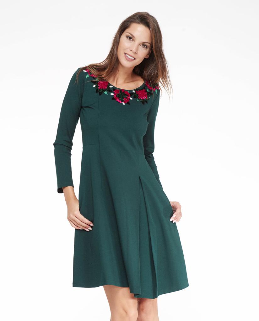 Uśmiechnięta kobieta w ciemnozielonej sukience z długim rękawem i haftowanymi czerwonymi różami wokół dekoltu na białym tle.