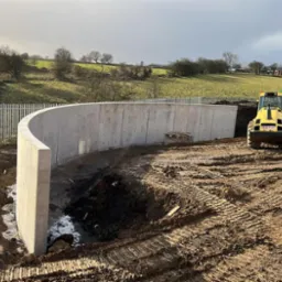 Żelbetowa konstrukcja w kształcie łuku w trakcie budowy na placu budowy z ciężkim sprzętem w tle, widok na wzgórza i częściowo zachmurzone niebo.
