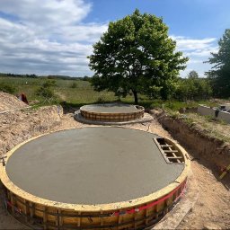 Fenix Construction - Dwa okrągłe, betonowe fundamenty w drewnianych szalunkach, w wykopie ziemnym na tle zieleni i błękitnego nieba z chmurami. Widoczne narzędzia i elementy konstrukcyjne.