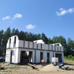 Fenix Construction - Surowy stan domu z białego pustaka, widoczna zaprawa, piasek, betoniarka i zbiornik na wodę. Budowa w toku, otoczenie leśne, błękitne niebo.