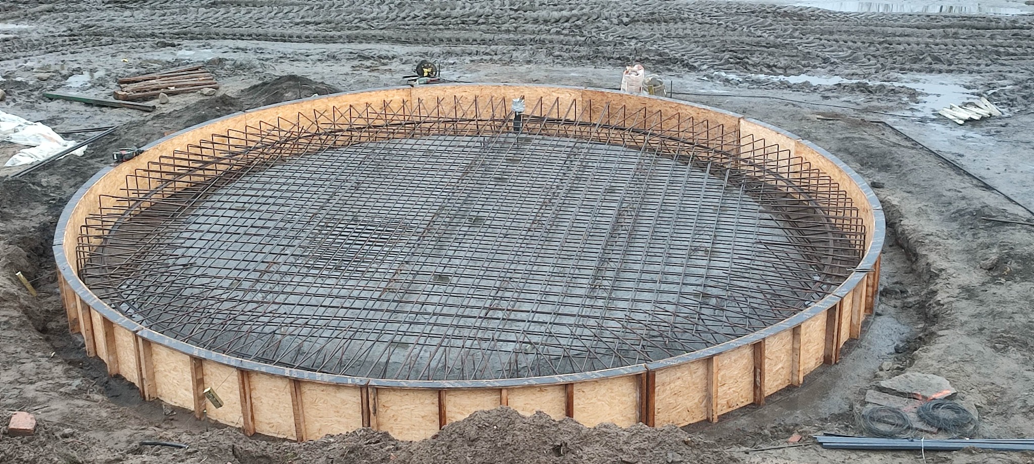 Okrągły szalunek z siatką zbrojeniową na przygotowanym podłożu, widoczne narzędzia i materiały budowlane w tle, ziemisty teren budowy.