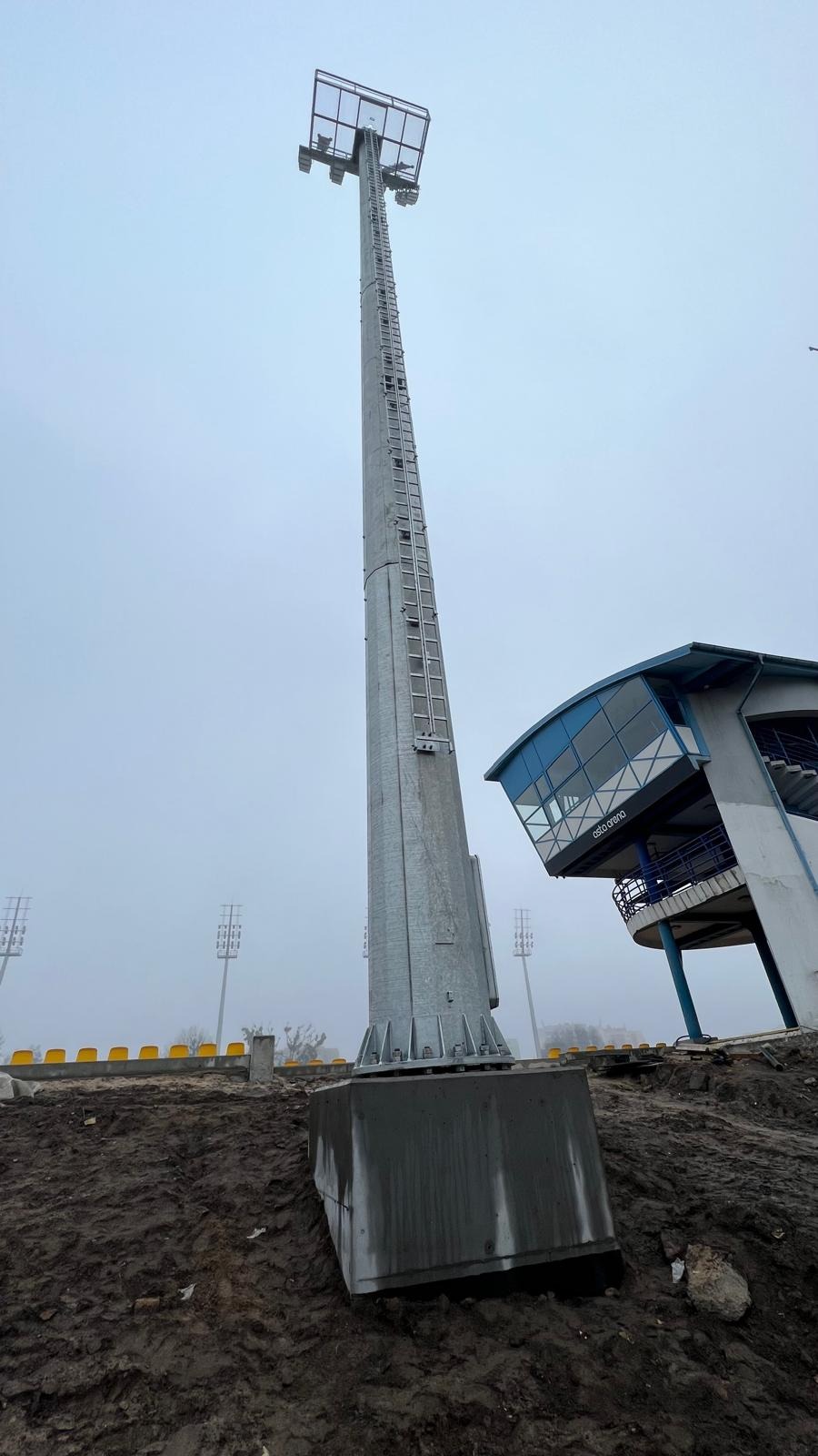 Stadion w mglisty dzień: wysoki maszt oświetleniowy z drabinką i betonową podstawą, obok budynek komentatorski z niebieskimi akcentami.