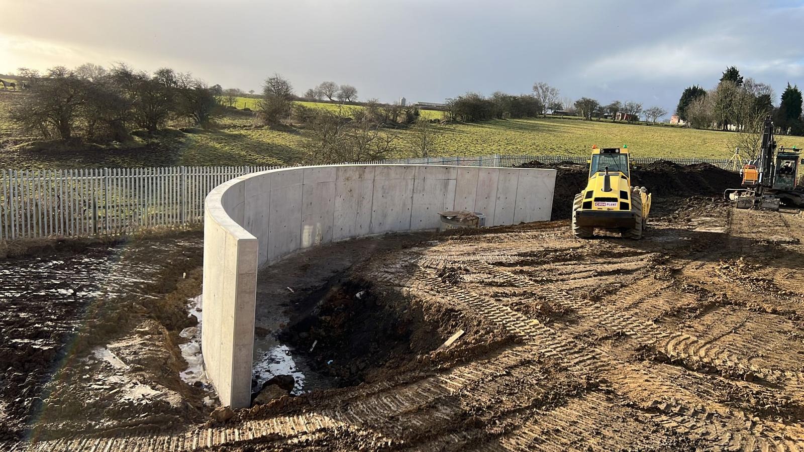 Żelbetowa konstrukcja w kształcie łuku w trakcie budowy na placu budowy z ciężkim sprzętem w tle, widok na wzgórza i częściowo zachmurzone niebo.