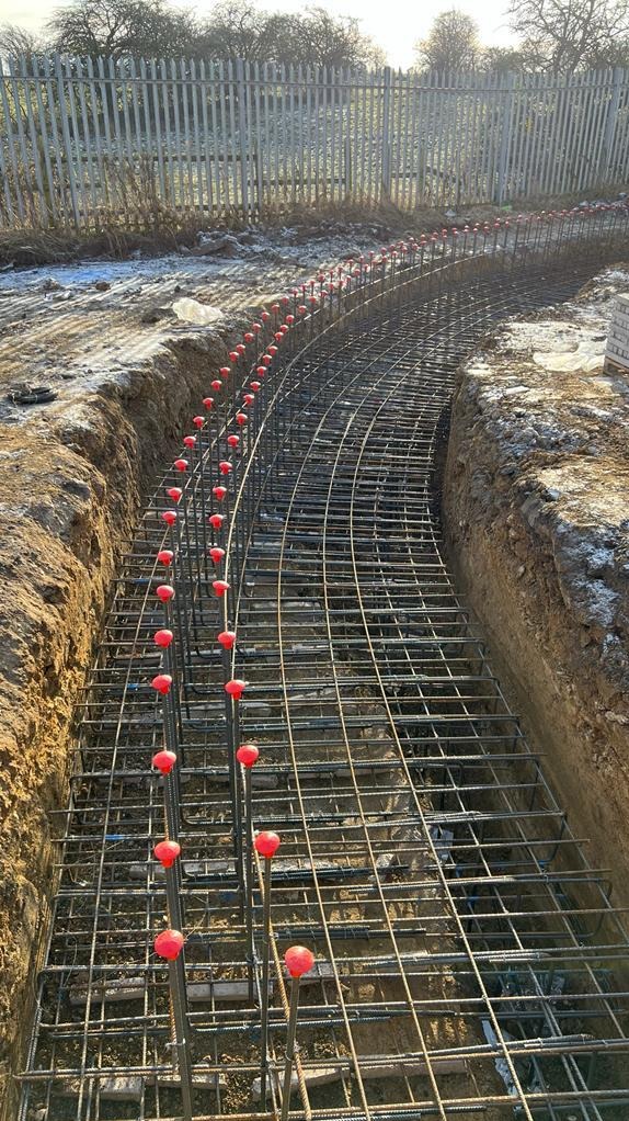 Zbrojenie fundamentu w wykopie, widoczne pręty zbrojeniowe z czerwonymi osłonami, ziemia z lekkim oszronieniem, w tle metalowe ogrodzenie.
