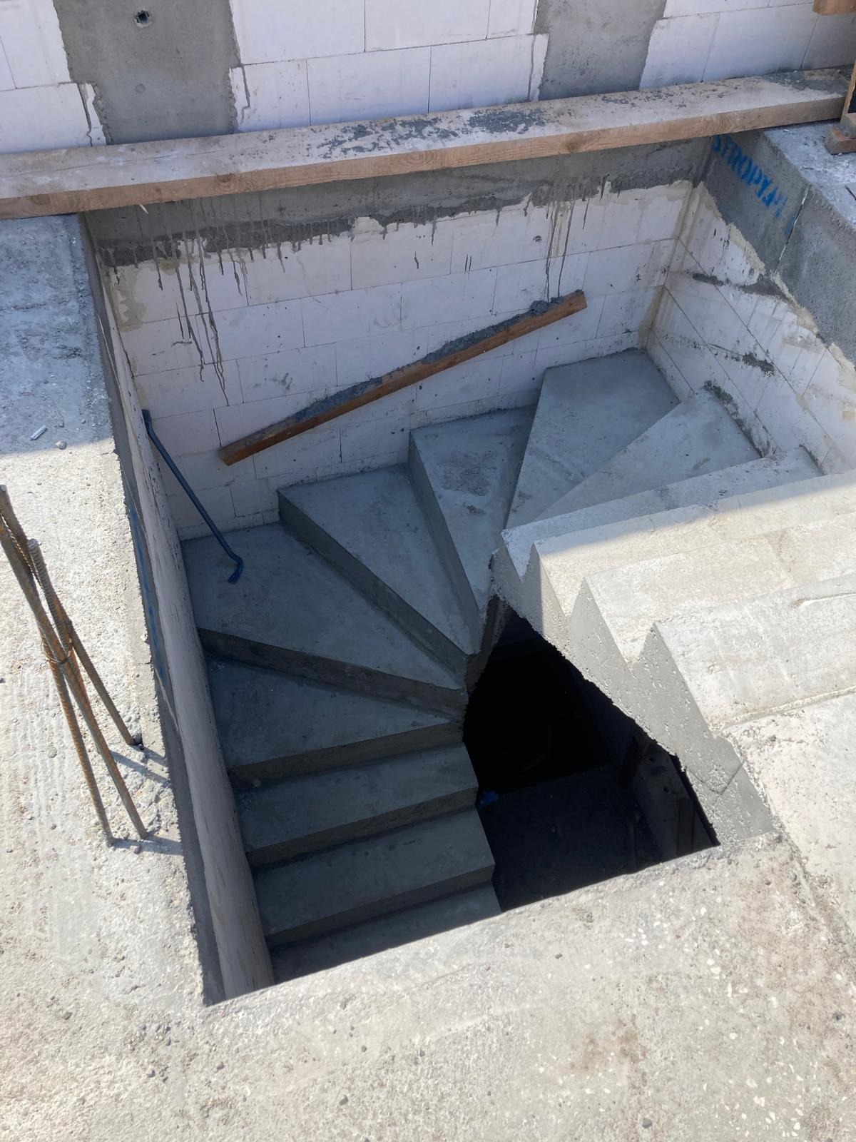 Wewnętrzne, spiralne schody z betonu w surowym stanie, widoczne z góry, z drewnianymi elementami szalunku i ścianami z bloczków betonowych.