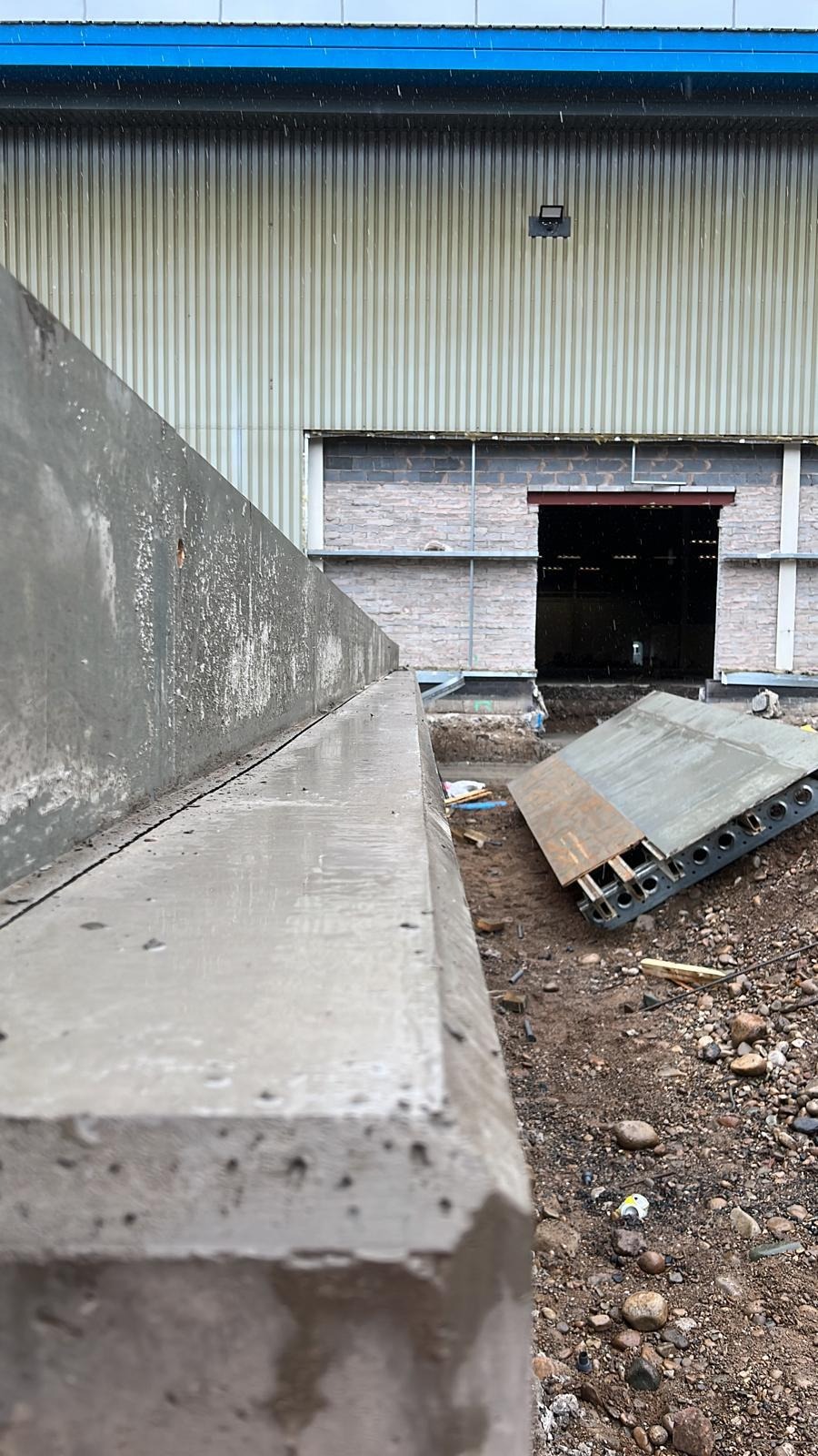 Surowy betonowy element konstrukcyjny w trakcie budowy, widoczne zbrojenie i niedokończone otoczenie placu budowy, tło stanowi hala przemysłowa.