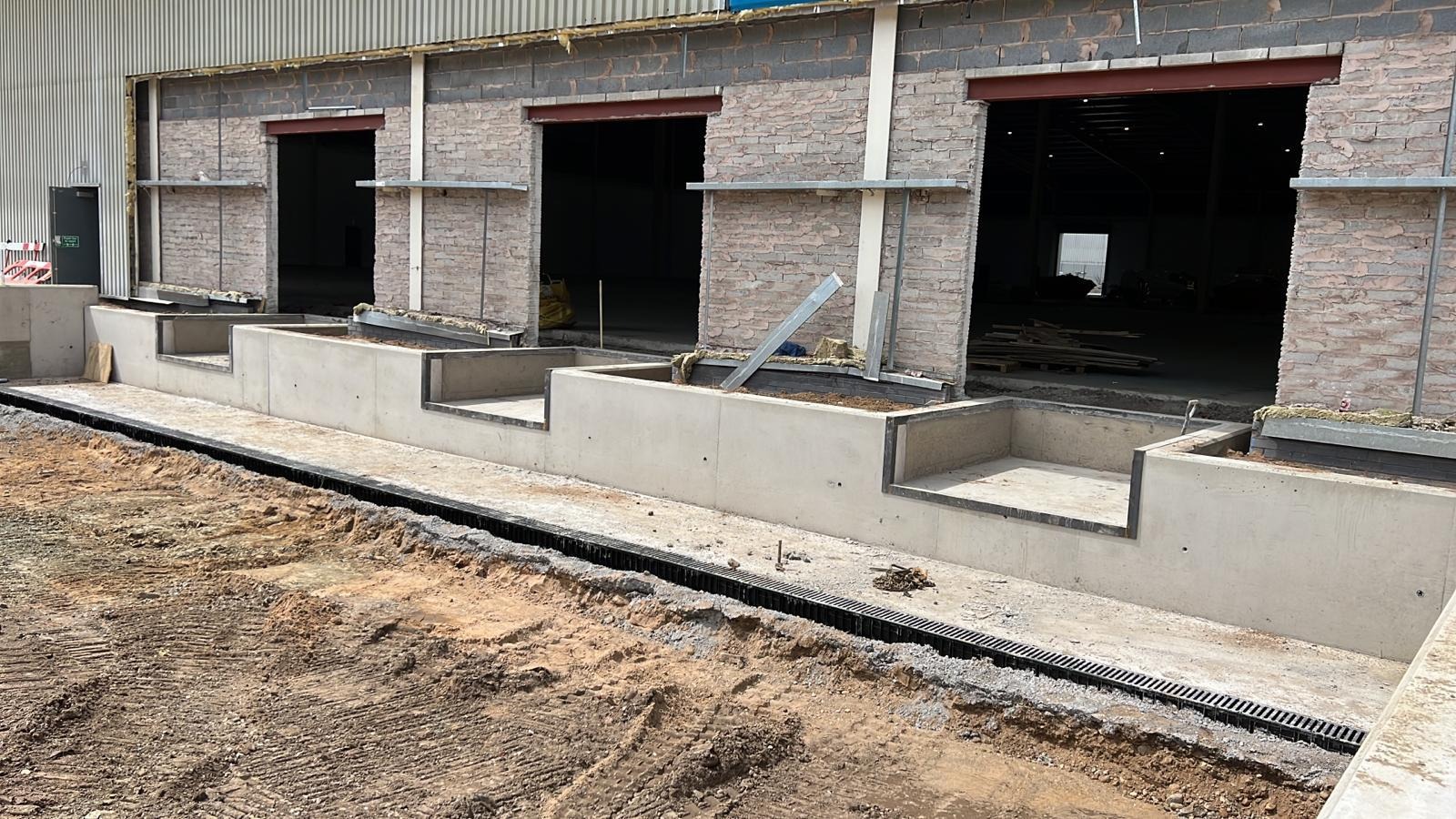 Surowe betonowe fundamenty pod budynek z trzema otworami wejściowymi, widoczny system odwadniający i przygotowany teren.