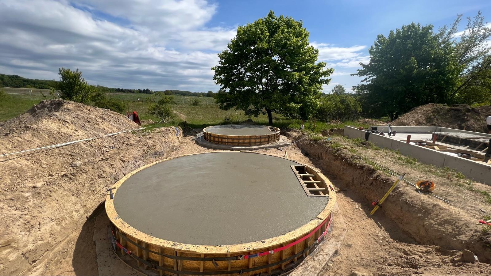Dwa okrągłe, betonowe fundamenty w drewnianych szalunkach, w wykopie ziemnym na tle zieleni i błękitnego nieba z chmurami. Widoczne narzędzia i elementy konstrukcyjne.
