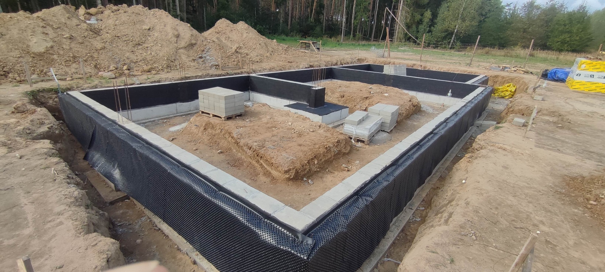Zabezpieczone fundamenty domu jednorodzinnego z hydroizolacją pionową i bloczkami betonowymi na palecie w wykopie. Widoczne zbrojenia i przygotowany teren pod budowę.