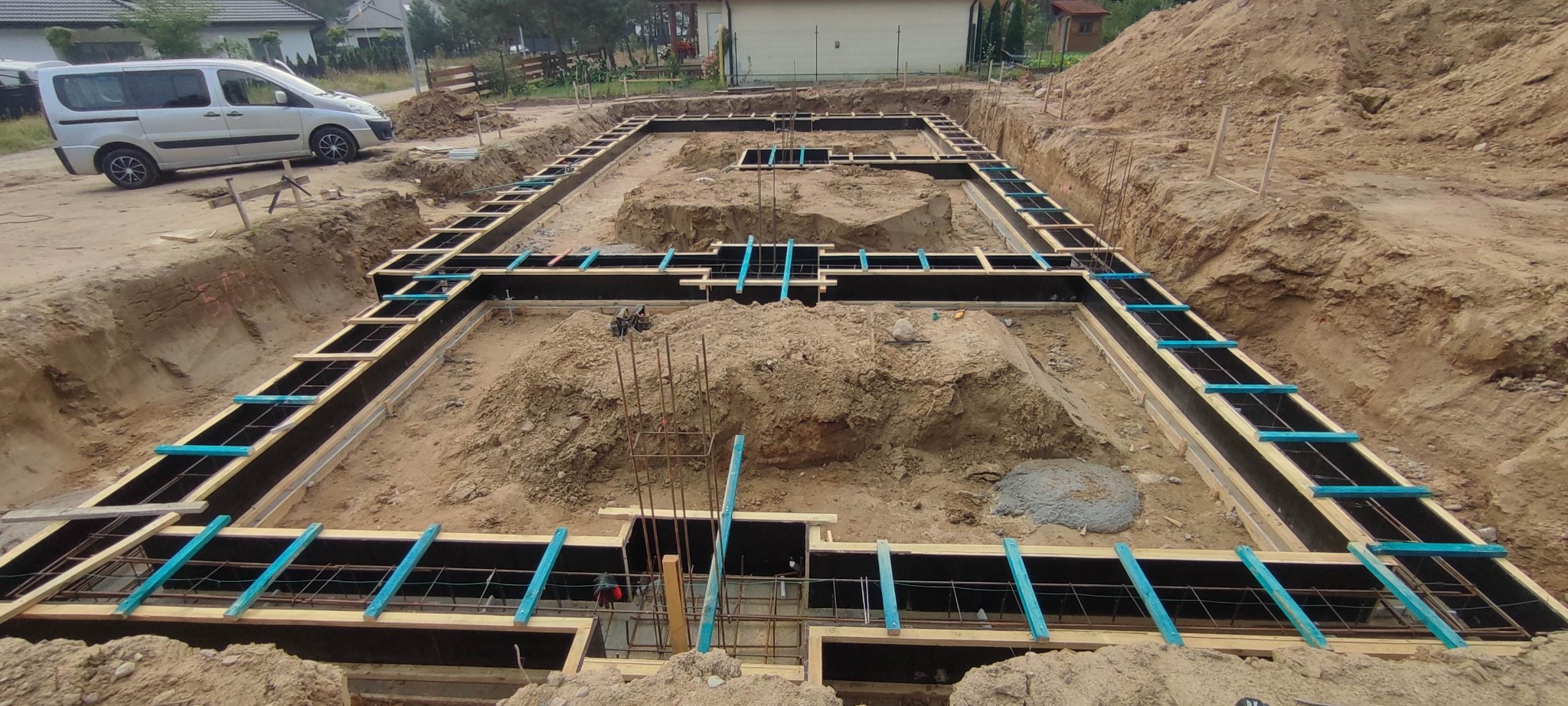 Przygotowanie szalunku pod fundamenty domu jednorodzinnego. Widoczne zbrojenie i wykop. W tle zaparkowany srebrny van.