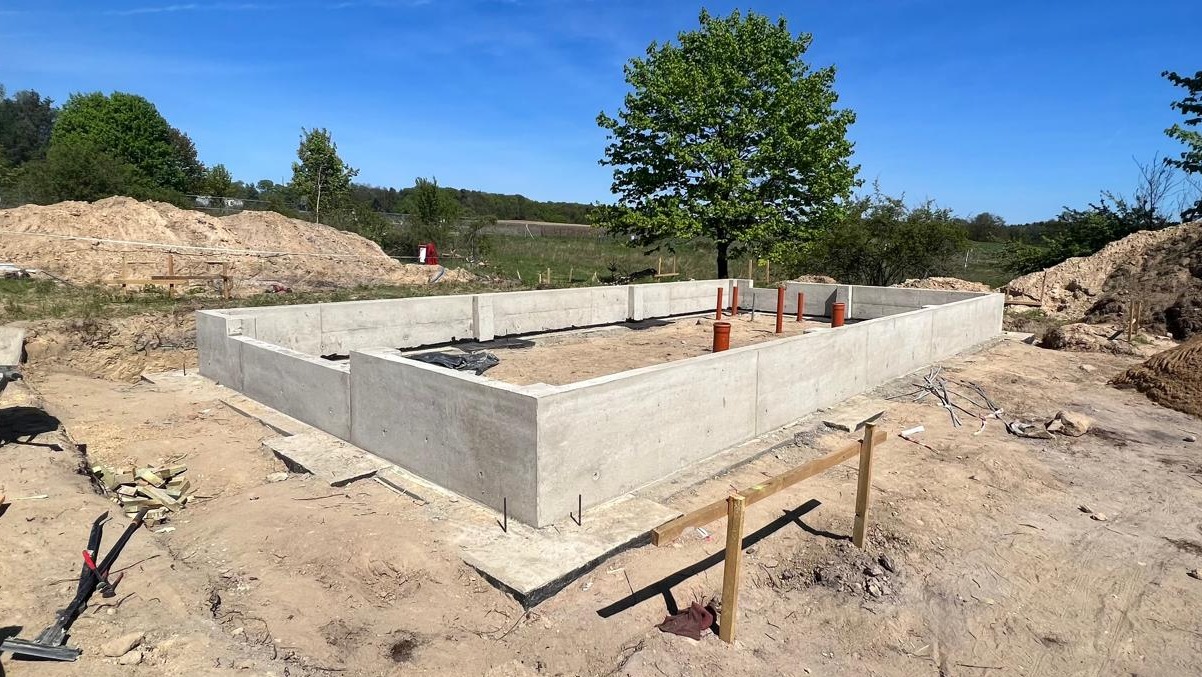 Zalane betonowe fundamenty domu jednorodzinnego z wystającymi rurami kanalizacyjnymi na tle zieleni i błękitnego nieba w słoneczny dzień.