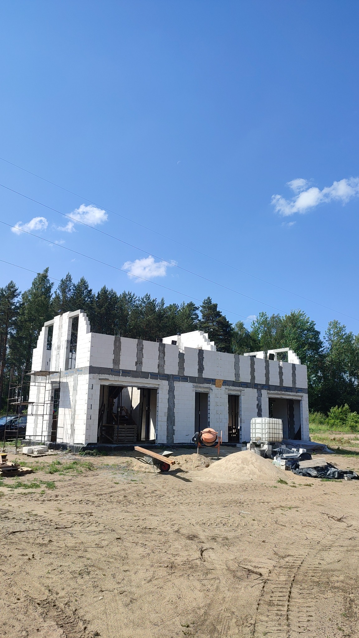 Surowy stan domu z białego pustaka, widoczna zaprawa, piasek, betoniarka i zbiornik na wodę. Budowa w toku, otoczenie leśne, błękitne niebo.