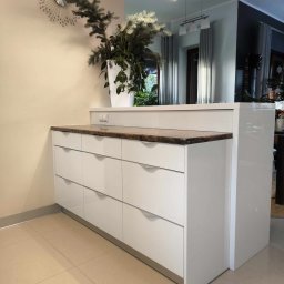 Meble ADAW Ojciec & Syn ADAM MAŁYSZKA - Biała komoda z sześcioma szufladami i blatem z brązowego kamienia, dekorowana roślinami w białej donicy, widok na salon z szarymi zasłonami