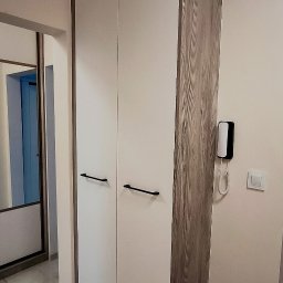 Meble ADAW Ojciec & Syn ADAM MAŁYSZKA - Dwudrzwiowa szafa na wymiar z białymi frontami i drewnopodobnym bokiem w przedpokoju. Widoczne czarne, podłużne uchwyty i fragment lustrzanej szafy przesuwnej.