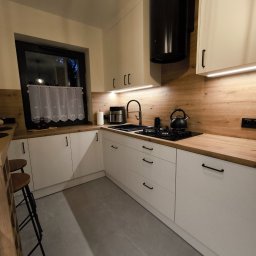 Meble ADAW Ojciec & Syn ADAM MAŁYSZKA - Nowoczesna kuchnia z białymi szafkami, drewnianym blatem i czarnymi akcentami, w tym okapem i armaturą. Uwagę przykuwa oświetlenie LED pod szafkami.