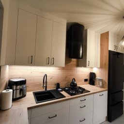 Meble ADAW Ojciec & Syn ADAM MAŁYSZKA - Nowoczesna kuchnia z białymi szafkami, czarnym zlewem i sprzętami AGD. Blat drewnopodobny, oświetlenie LED pod szafkami. Minimalistyczny design z czarnymi uchwytami.