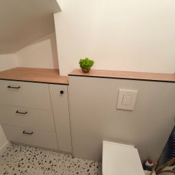 Meble ADAW Ojciec & Syn ADAM MAŁYSZKA - Biała szafka z trzema szufladami i drewnianym blatem w łazience na poddaszu. Na blacie dekoracyjna roślina. Podłoga w stylu lastryko.