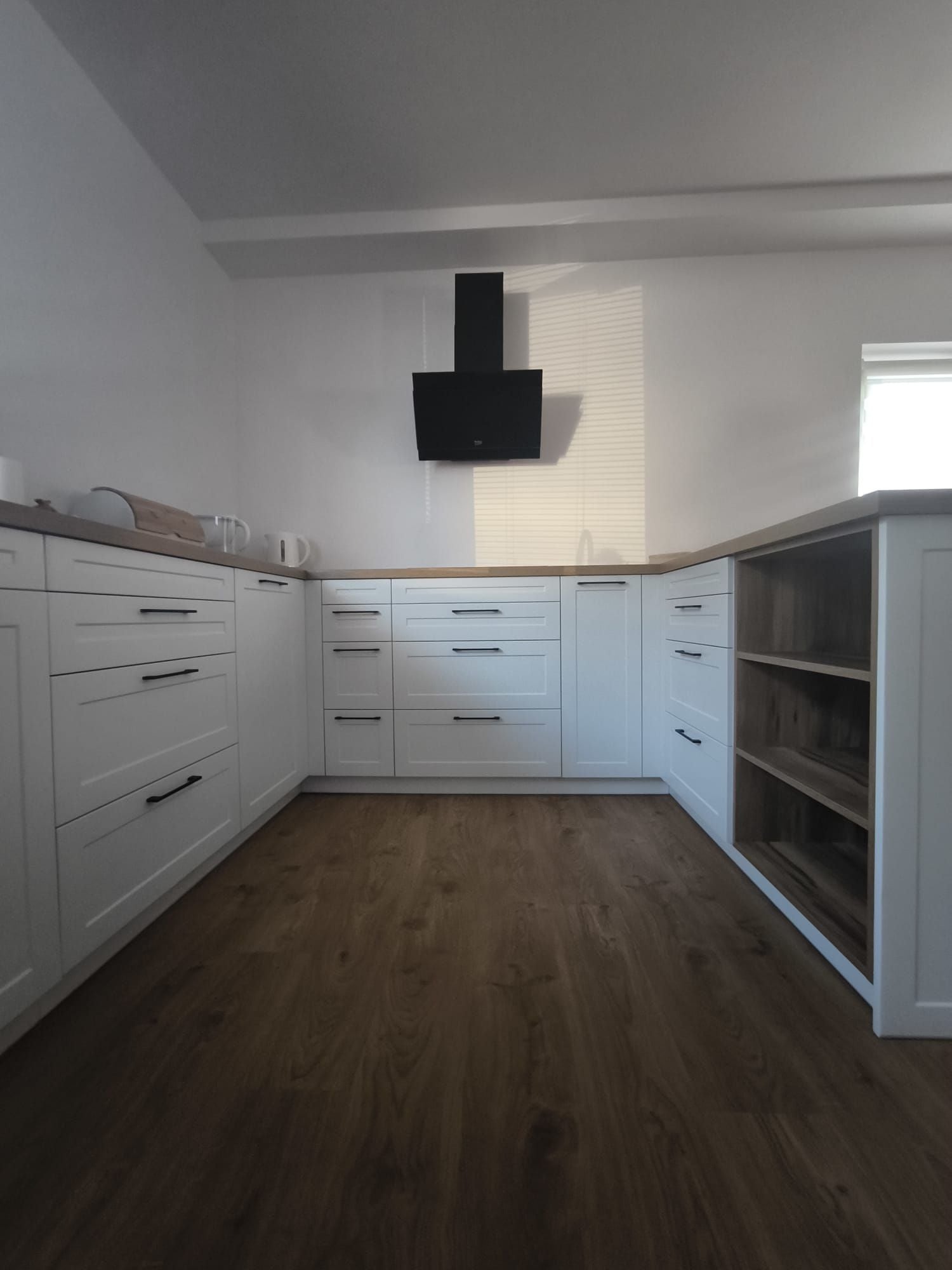 Nowoczesna biała kuchnia z drewnianym blatem i czarnym okapem, widok na przestronne wnętrze i drewnianą podłogę.