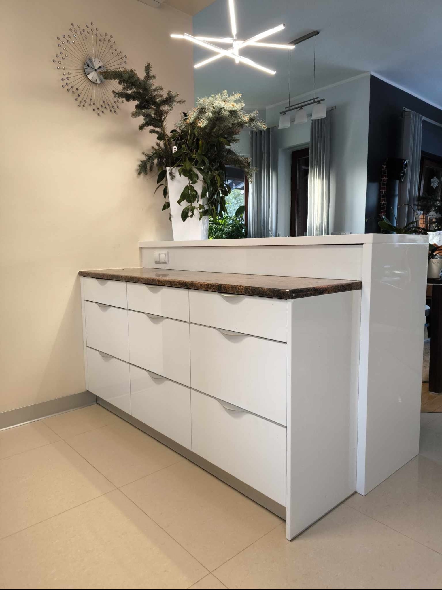 Biała komoda z sześcioma szufladami i blatem z brązowego kamienia, dekorowana roślinami w białej donicy, widok na salon z szarymi zasłonami