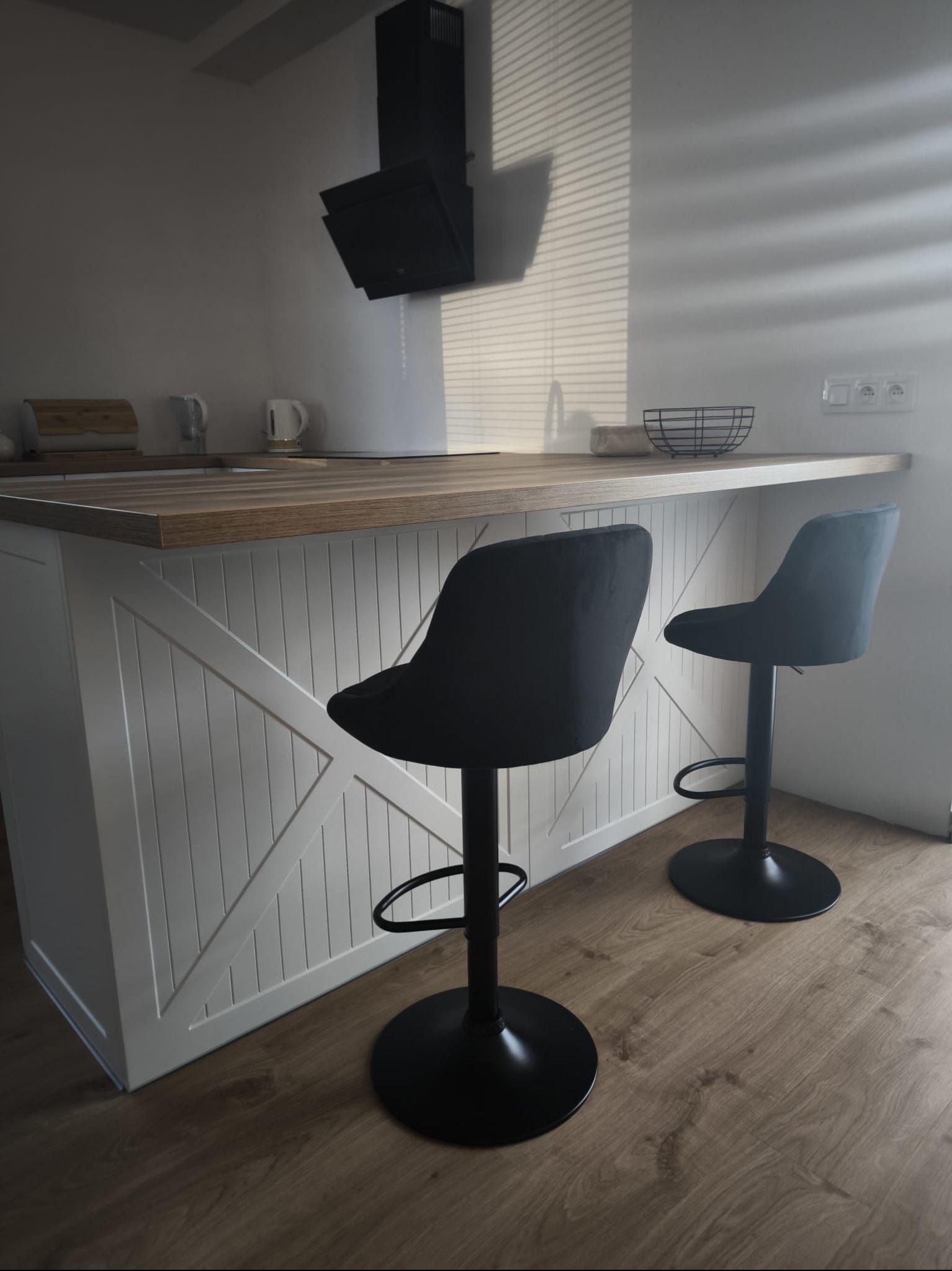 Nowoczesna wyspa kuchenna z drewnianym blatem i białymi frontami w stylu rustykalnym, uzupełniona dwoma czarnymi hokerami w Gruszczynie, oświetlona naturalnym światłem zza żaluzji.
