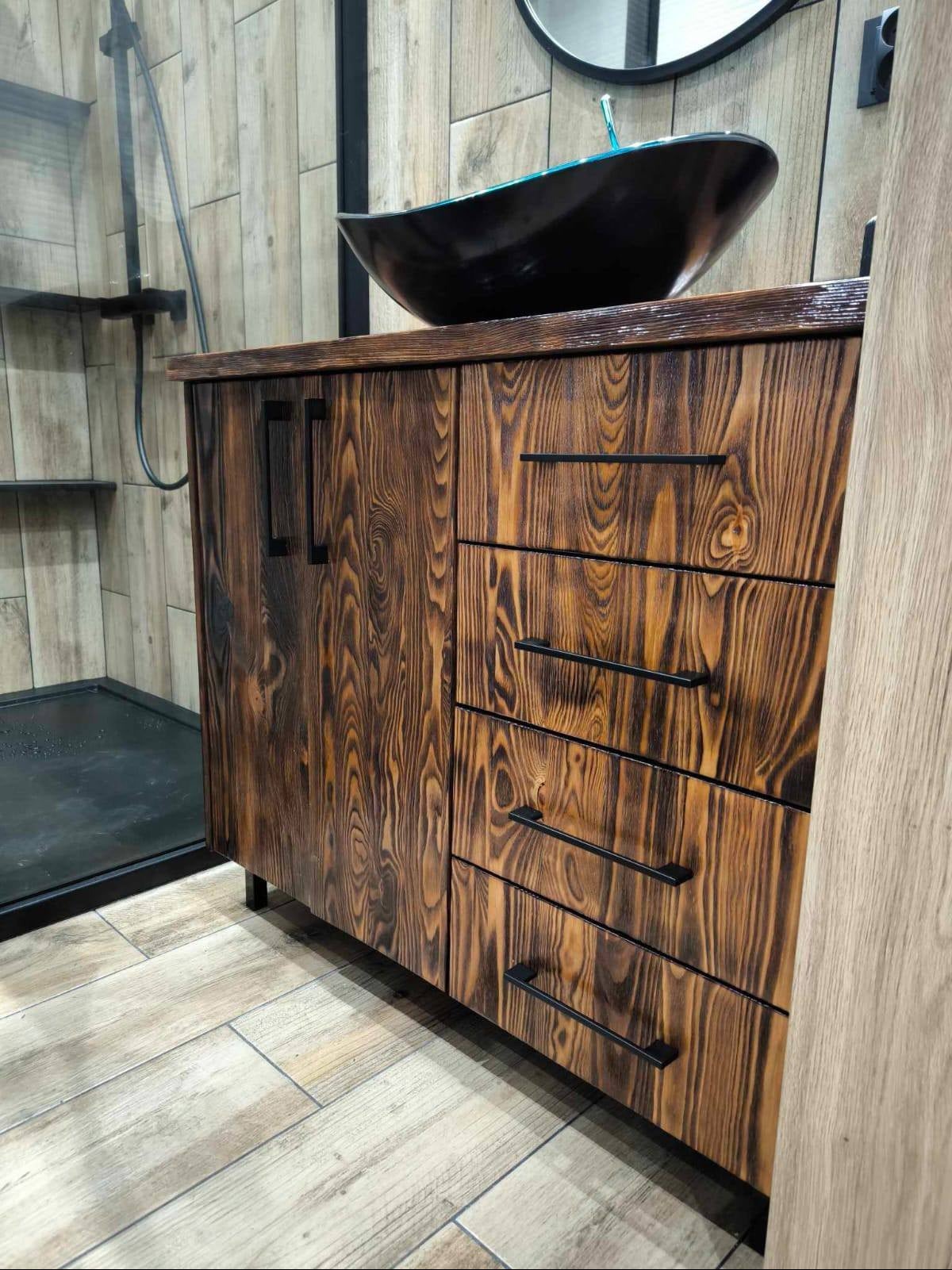 Brązowa szafka łazienkowa z palonego drewna z czarnymi uchwytami szuflad i czarną umywalką na blacie, obok kabina prysznicowa z czarną armaturą.