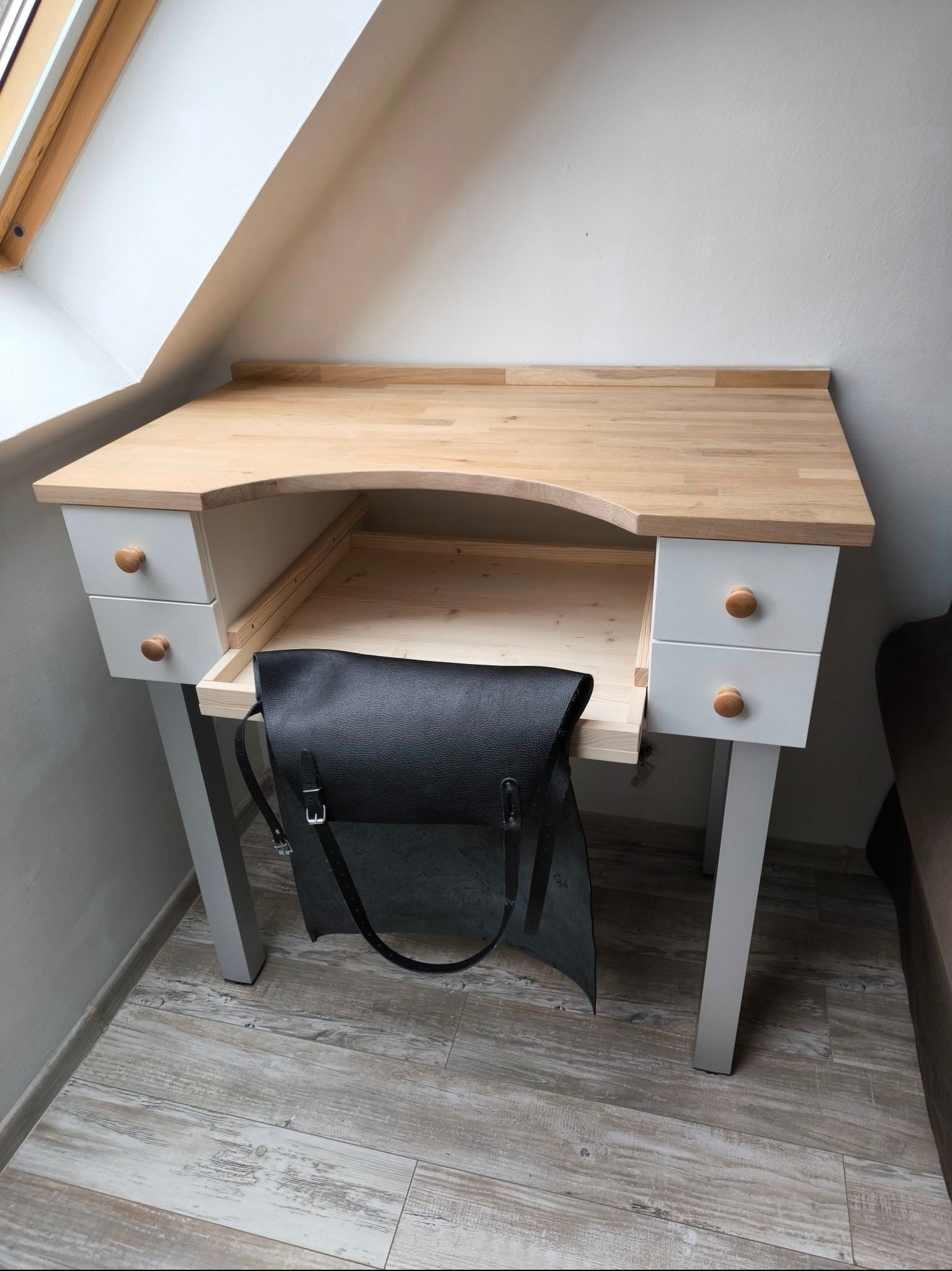 Drewniane biurko z jasnym blatem, białymi szufladami i okrągłymi uchwytami, z czarną torbą zwisającą z wysuniętej szuflady, ustawione w pokoju na poddaszu z oknem dachowym.