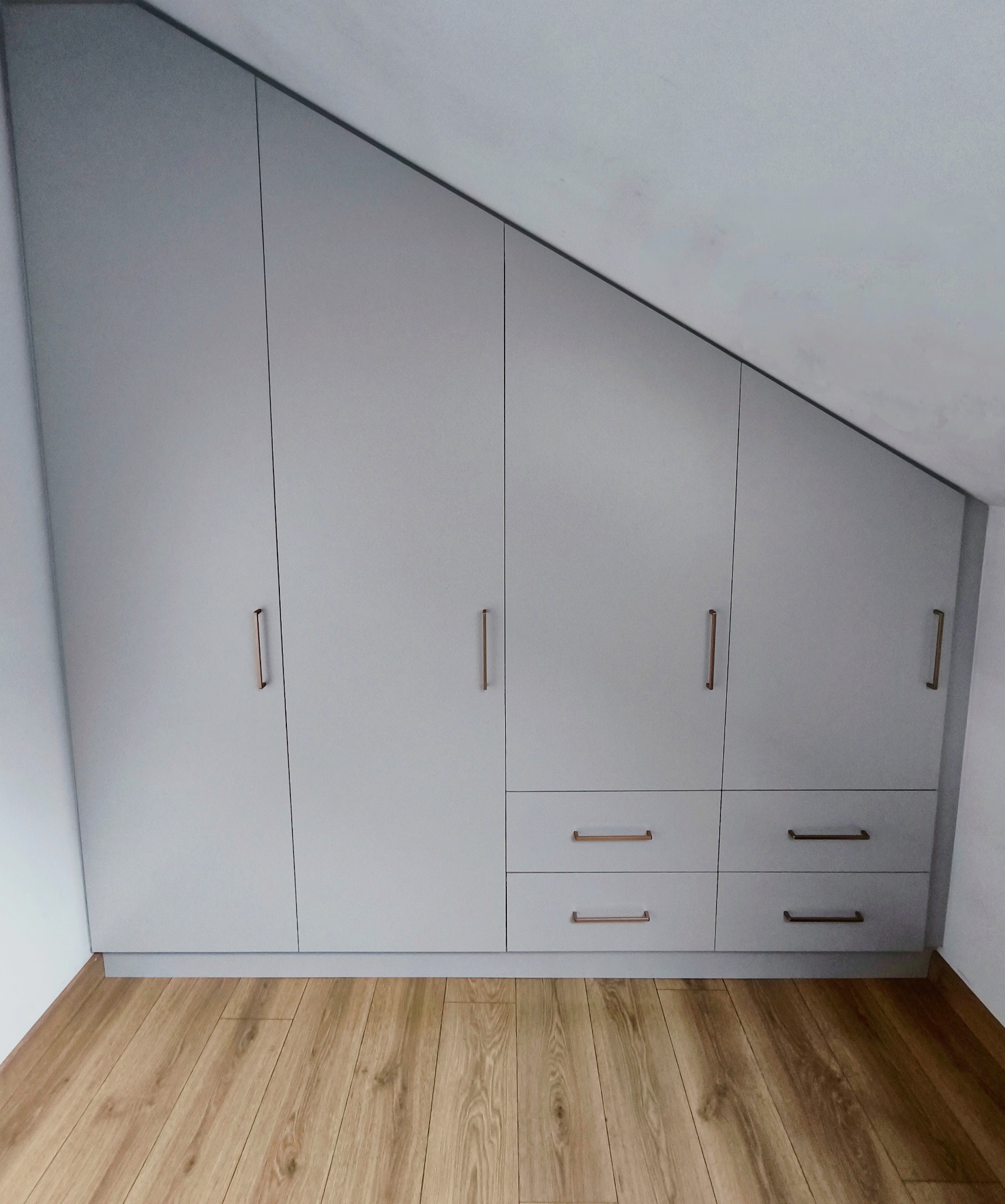 Jasnoszara szafa wnękowa na poddaszu z czterema drzwiami i dwoma rzędami szuflad, z podłużnymi, miedzianymi uchwytami, na drewnianej podłodze.