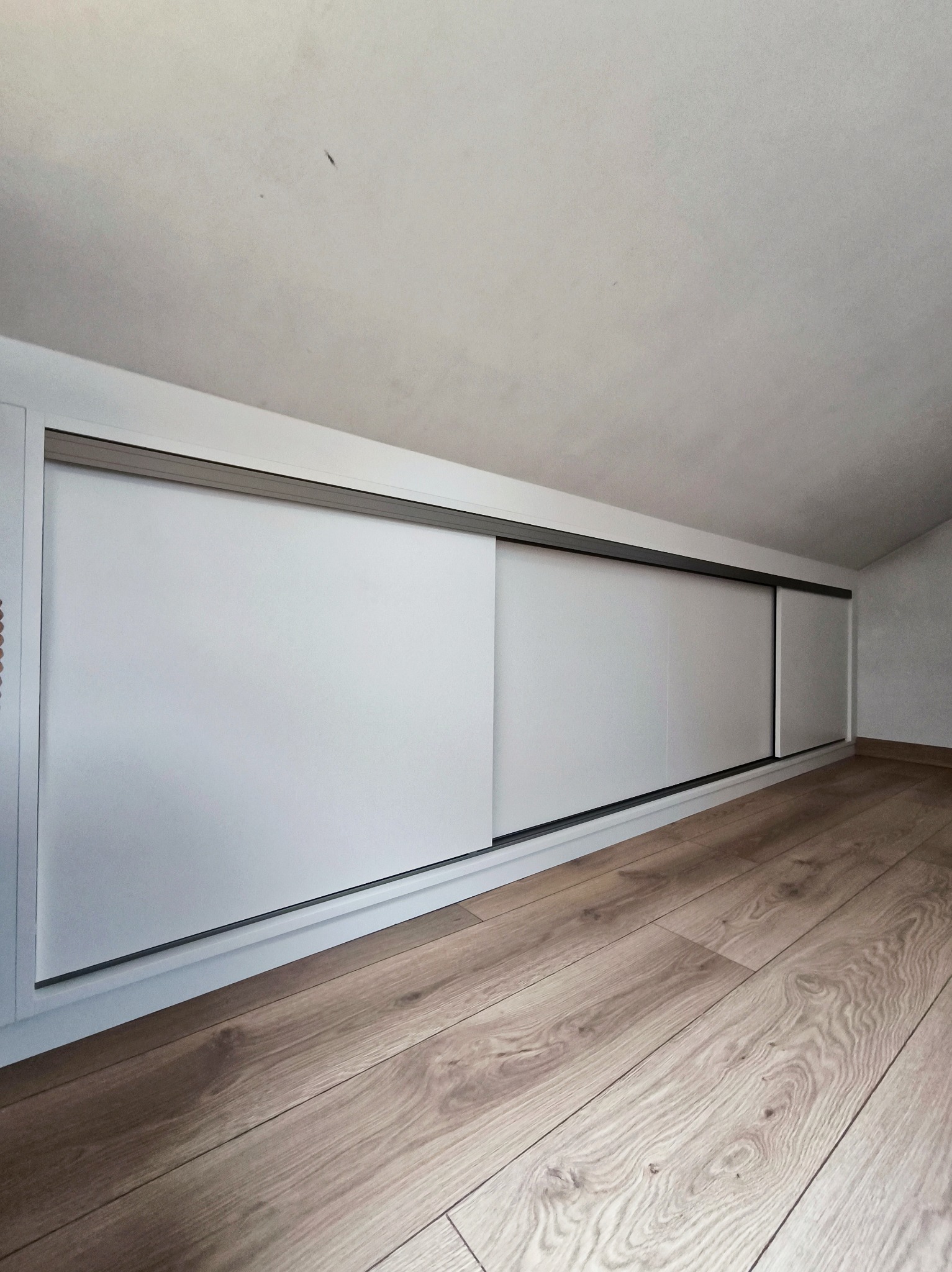 Biała szafa wnękowa z przesuwnymi drzwiami, zamontowana na poddaszu ze skośnym sufitem i drewnianą podłogą. Minimalistyczny design.